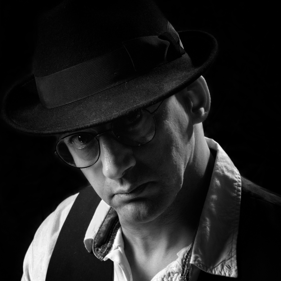 Kamel le photographe - KBFotoArt Black-and-white portrait of a man wearing a fedora, glasses, and a vest, looking downward with moody lighting.