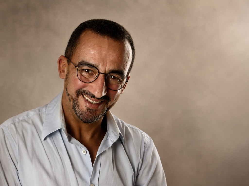 Portrait of a smiling man with glasses and stubble, wearing a light blue button-down shirt, against a neutral beige studio backdrop.